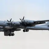 Grauer Tupolew Tu-142 mit vier Propellern im Flug, Fahrwerk ausgefahren, vor hellem Himmel.
