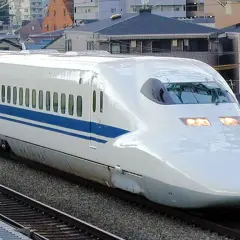 Ein weißer Shinkansen mit blauen Streifen rast auf den Gleisen entlang. Die **Shinkansen Geschwindigkeit** ist beeindruckend, während er an Häusern vorbeifährt.