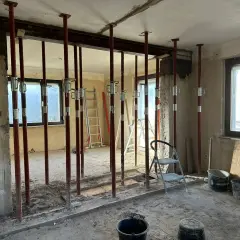 Baustelle mit vielen roten Stützen, die einen schweren **Eisenstrahl** tragen. Werkzeuge und Eimer liegen herum.