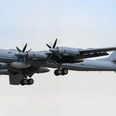 Grauer Tupolew Tu-142 mit vier Propellern im Flug, Fahrwerk ausgefahren, vor hellem Himmel.