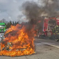 Ein **Elektroauto brennt** lichterloh auf der Autobahn. Feuerwehrleute versuchen, die Flammen zu löschen, während der Verkehr stockt.