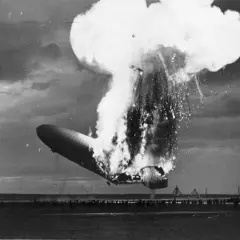 Ein Zeppelin stürzt brennend ab, eine riesige Explosion reißt ihn auseinander. Zuschauer stehen fassungslos am Boden.