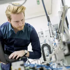 Ingenieur mit Laptop vor komplexer Maschine. So machen Ingenieure Technik greifbar und entwickeln Lösungen.