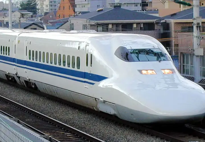 Ein weißer Shinkansen mit blauen Streifen rast auf den Gleisen entlang. Die **Shinkansen Geschwindigkeit** ist beeindruckend, während er an Häusern vorbeifährt.