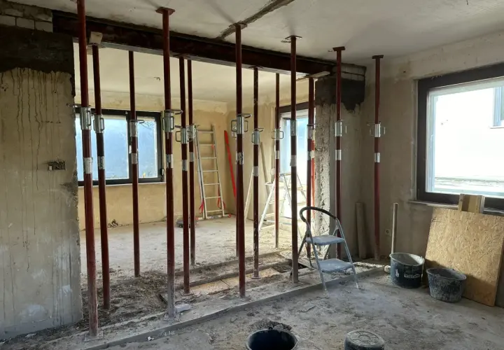 Baustelle mit vielen roten Stützen, die einen schweren **Eisenstrahl** tragen. Werkzeuge und Eimer liegen herum.