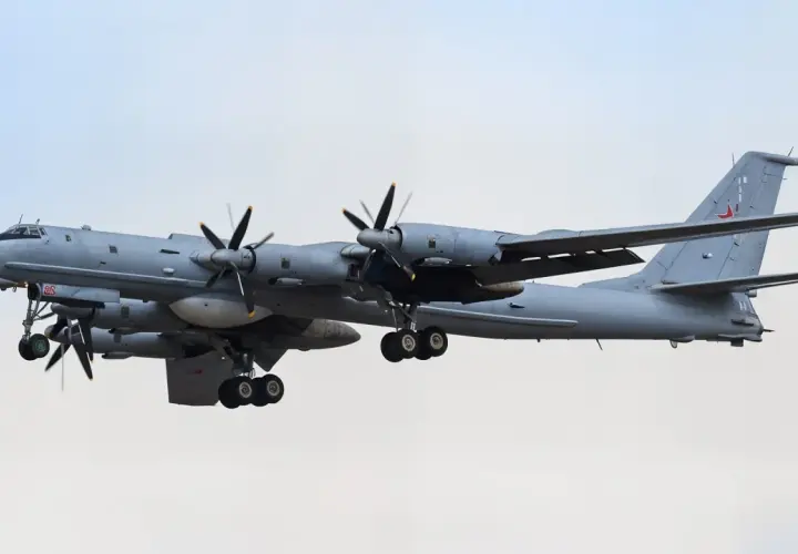 Grauer Tupolew Tu-142 mit vier Propellern im Flug, Fahrwerk ausgefahren, vor hellem Himmel.