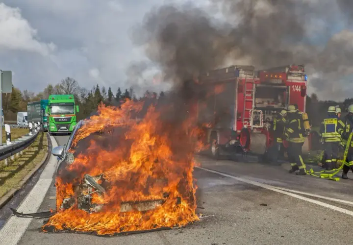 Ein **Elektroauto brennt** lichterloh auf der Autobahn. Feuerwehrleute versuchen, die Flammen zu löschen, während der Verkehr stockt.