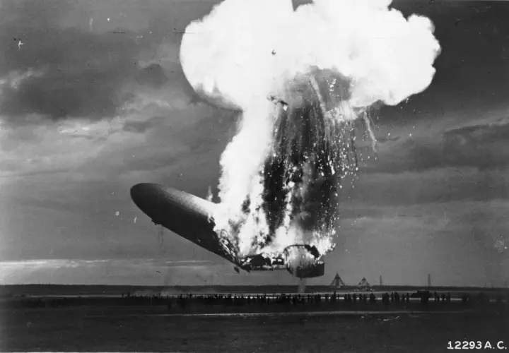 Ein Zeppelin stürzt brennend ab, eine riesige Explosion reißt ihn auseinander. Zuschauer stehen fassungslos am Boden.