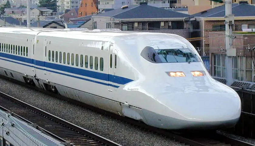 Ein weißer Shinkansen mit blauen Streifen rast auf den Gleisen entlang. Die **Shinkansen Geschwindigkeit** ist beeindruckend, während er an Häusern vorbeifährt.