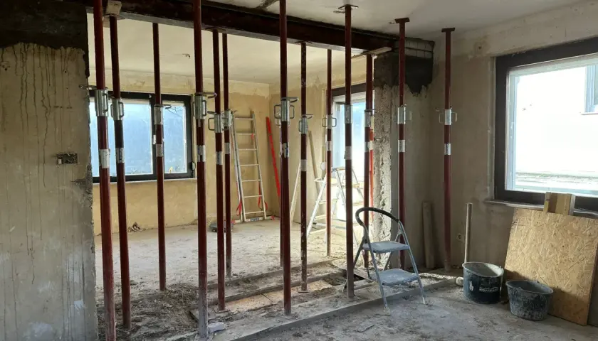 Baustelle mit vielen roten Stützen, die einen schweren **Eisenstrahl** tragen. Werkzeuge und Eimer liegen herum.