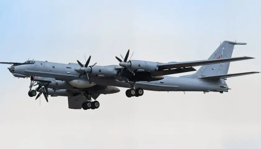 Grauer Tupolew Tu-142 mit vier Propellern im Flug, Fahrwerk ausgefahren, vor hellem Himmel.