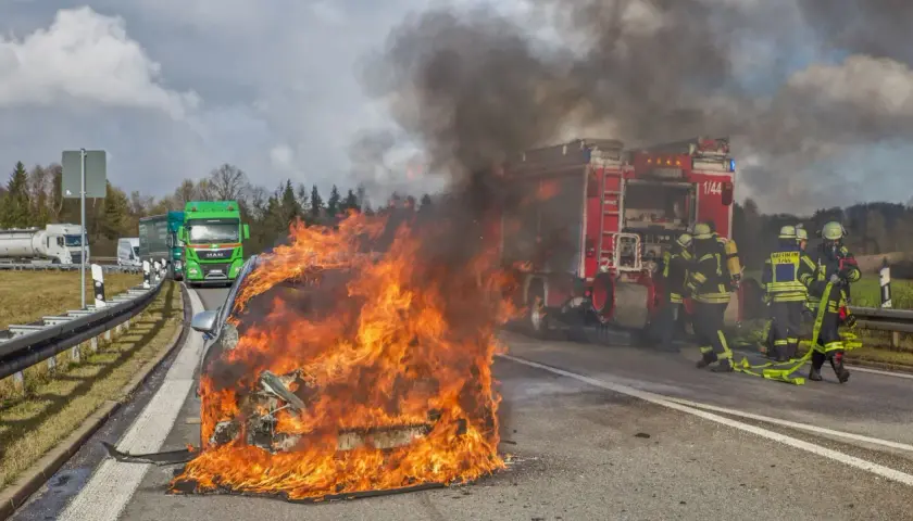 Ein **Elektroauto brennt** lichterloh auf der Autobahn. Feuerwehrleute versuchen, die Flammen zu löschen, während der Verkehr stockt.