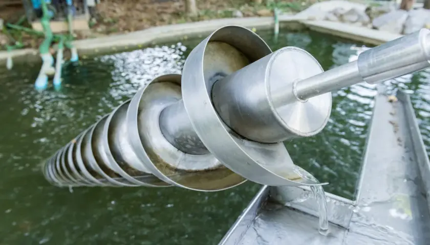 Eine Archimedes-Schraube hebt Wasser aus einem Teich. Das Wasser fließt über die spiralförmigen Windungen und wird nach oben transportiert.