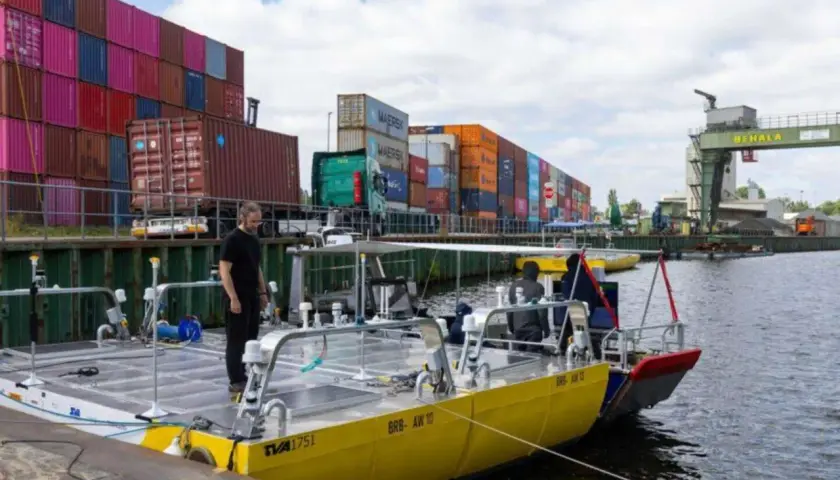 Ein Mann steht auf einem gelben Katamaran, der wie eine schwimmende Stadt aussieht, mit Frachtcontainern im Hintergrund.