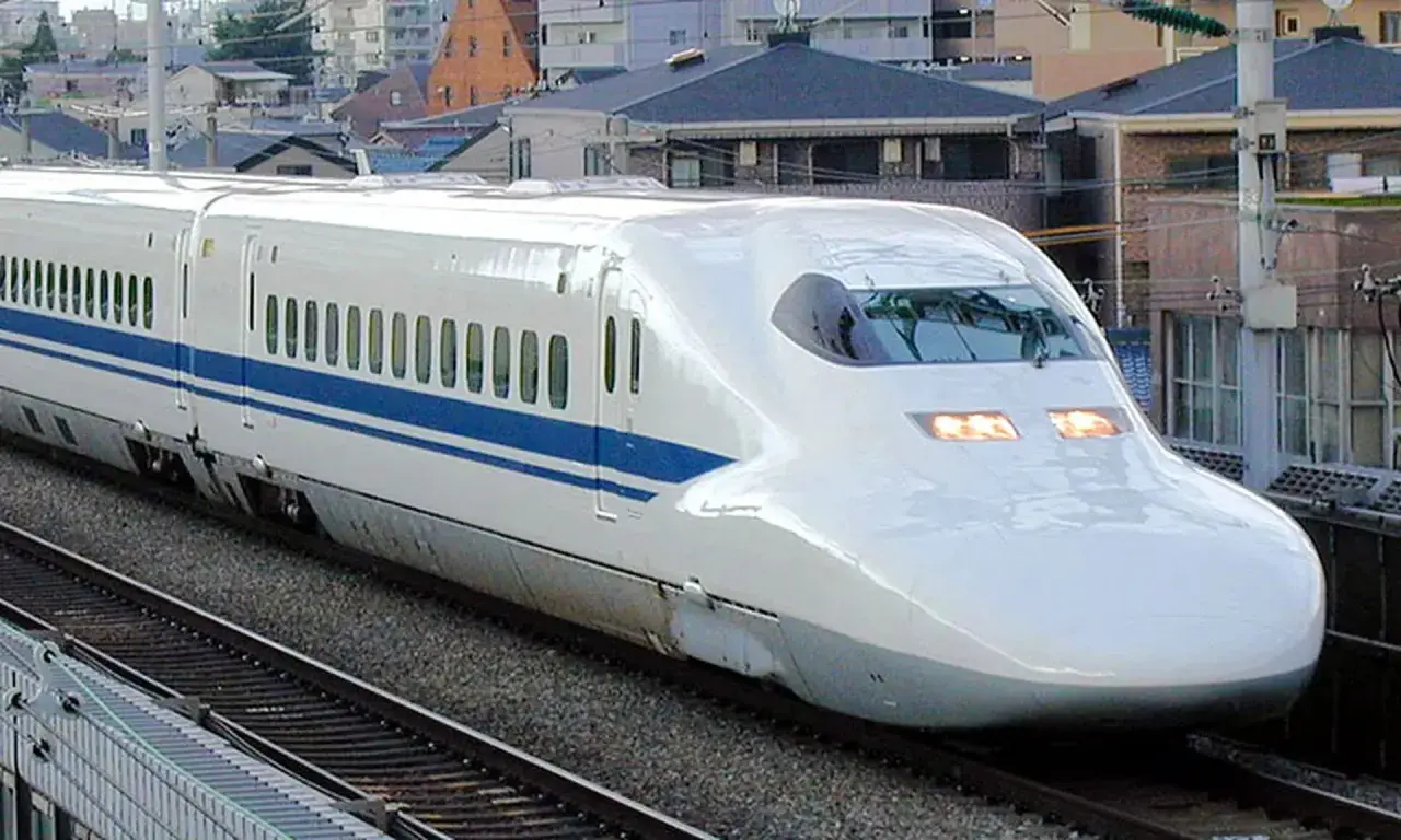 Ein weißer Shinkansen mit blauen Streifen rast auf den Gleisen entlang. Die **Shinkansen Geschwindigkeit** ist beeindruckend, während er an Häusern vorbeifährt.