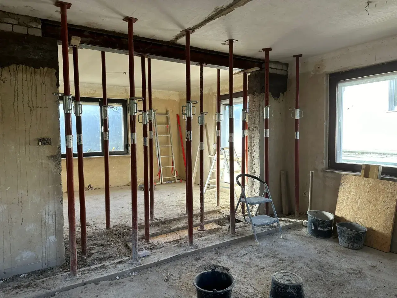 Baustelle mit vielen roten Stützen, die einen schweren **Eisenstrahl** tragen. Werkzeuge und Eimer liegen herum.