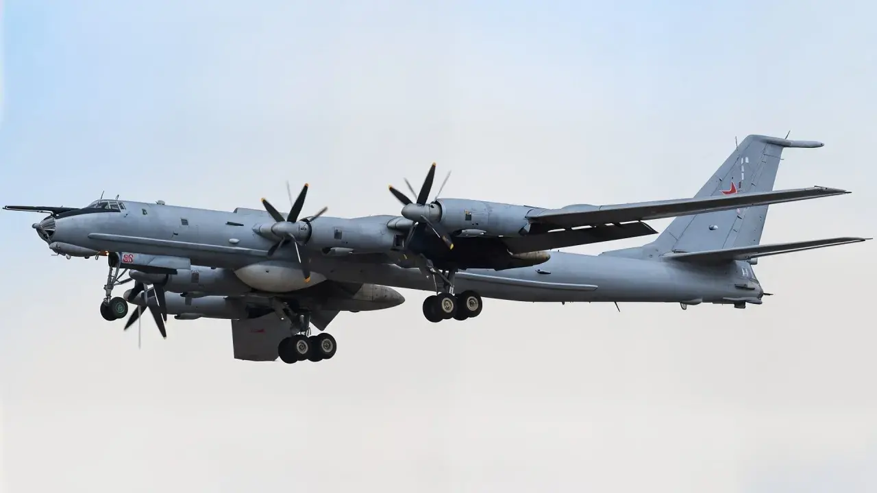 Grauer Tupolew Tu-142 mit vier Propellern im Flug, Fahrwerk ausgefahren, vor hellem Himmel.
