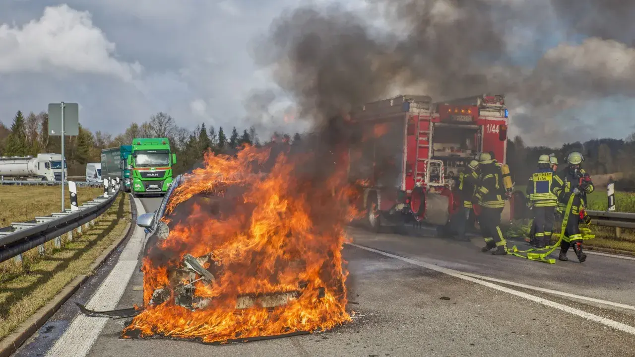 Ein **Elektroauto brennt** lichterloh auf der Autobahn. Feuerwehrleute versuchen, die Flammen zu löschen, während der Verkehr stockt.