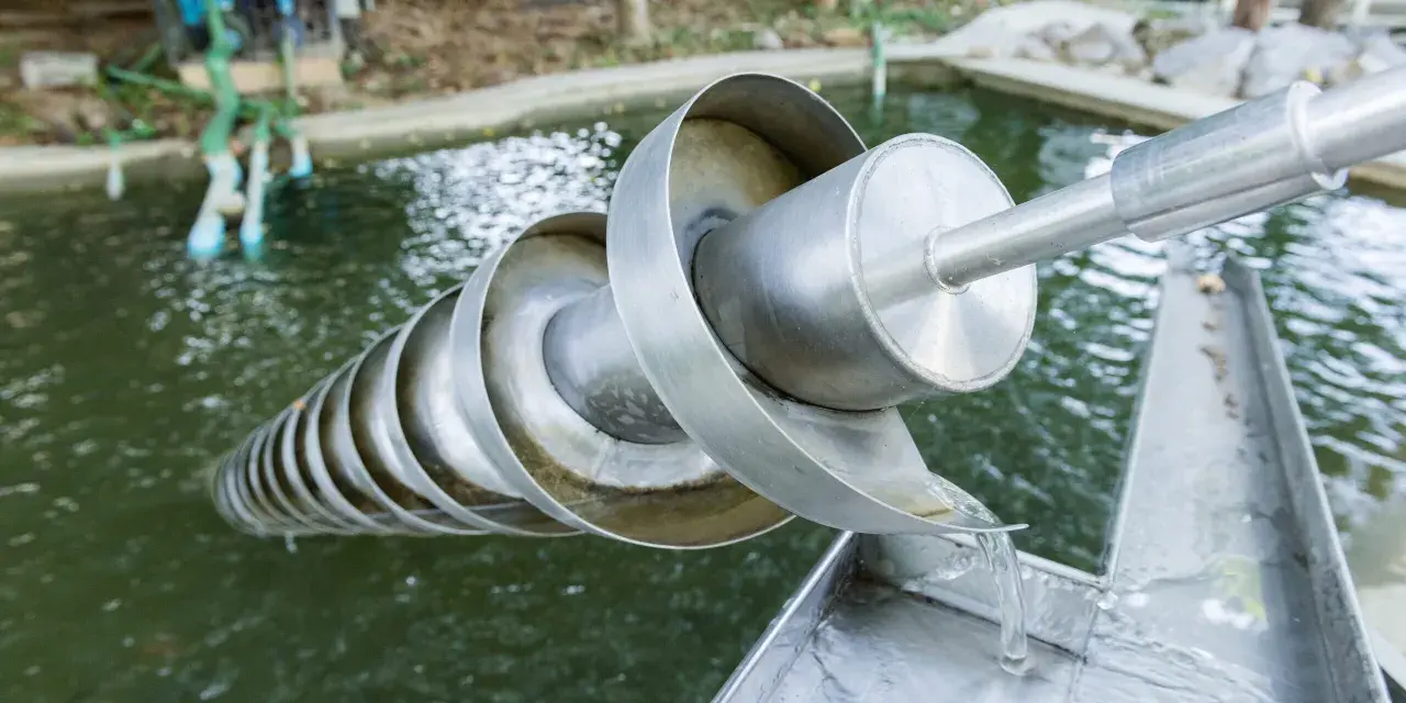Eine Archimedes-Schraube hebt Wasser aus einem Teich. Das Wasser fließt über die spiralförmigen Windungen und wird nach oben transportiert.