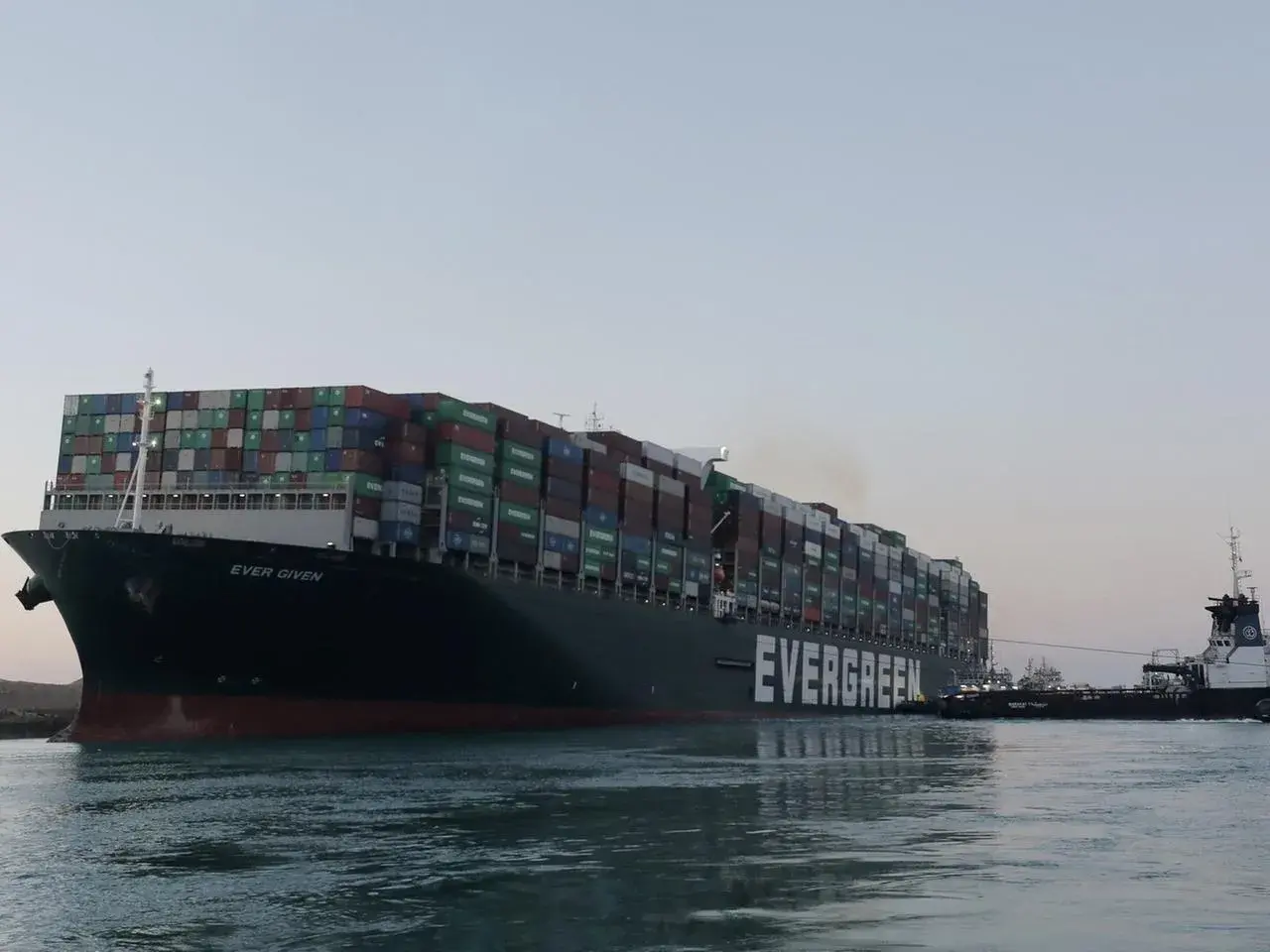 Ein riesiges Evergreen-Containerschiff, die "Ever Given", wird von einem Schlepper durch ruhiges Wasser gezogen.