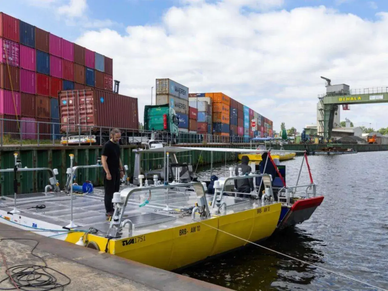 Ein Mann steht auf einem gelben Katamaran, der wie eine schwimmende Stadt aussieht, mit Frachtcontainern im Hintergrund.