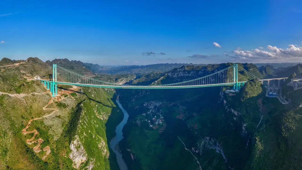 Die Huajiang Grand Canyon Bridge &uuml;berspannt eine tiefe Schlucht mit einem Fluss. Majest&auml;tische Berge und gr&uuml;ne H&auml;nge umgeben die beeindruckende Br&uuml;cke.