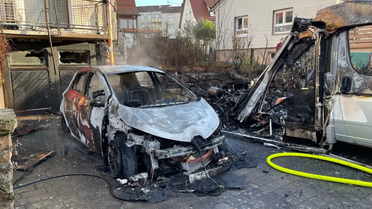 Ein ausgebranntes Elektroauto brennt neben einem zerstörten Wohnmobil. Rauch steigt auf, und ein gelber Schlauch liegt auf dem Boden.