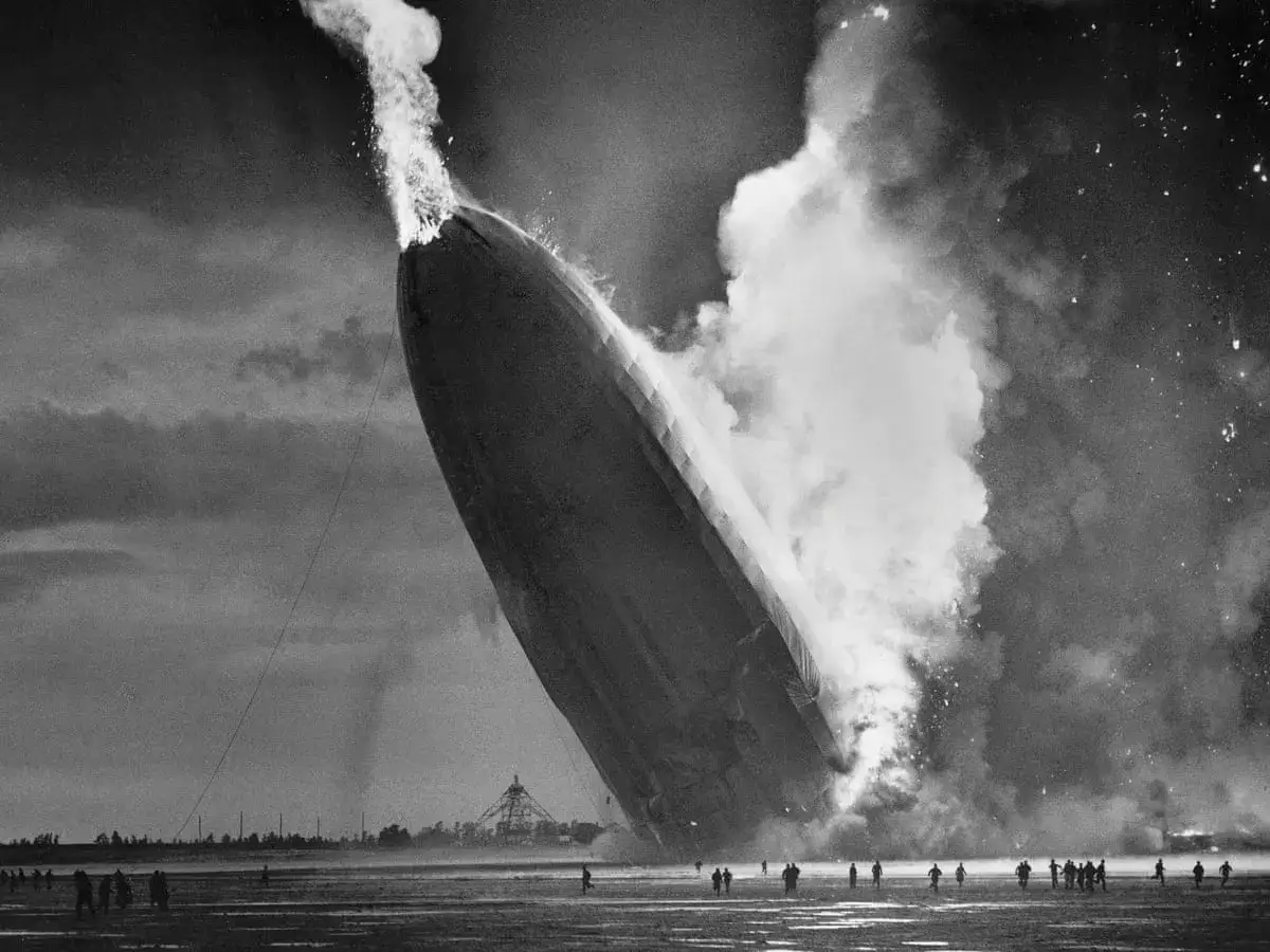 Das Luftschiff Hindenburg brennt und stürzt ab. Rauch und Flammen steigen auf, Menschen fliehen.