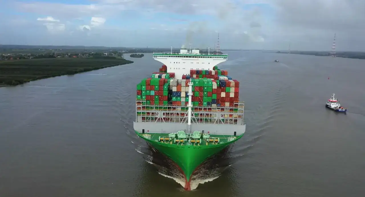 Ein riesiges Containerschiff, ein everalot Schiff, f&auml;hrt auf einem breiten Fluss. Im Hintergrund sind T&uuml;rme und eine kleine Stadt zu sehen.