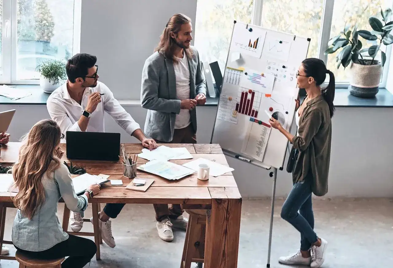 Ingenieure diskutieren bei einer Pr&auml;sentation. Eine Frau erkl&auml;rt Diagramme auf einem Whiteboard, w&auml;hrend ihre Kollegen aufmerksam zuh&ouml;ren.