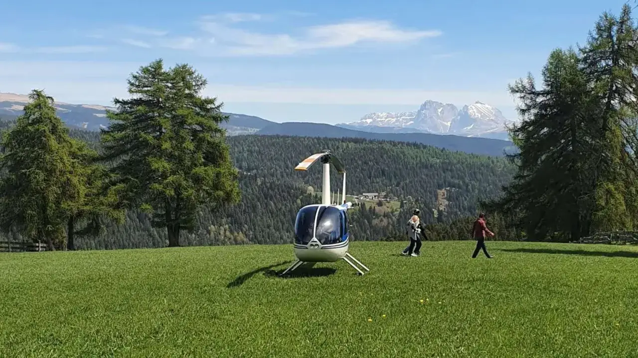 Ein kleiner Hubschrauber steht auf einer grünen Wiese. Im Hintergrund bewaldete Berge und schneebedeckte Gipfel. Man fragt sich, wie schnell fliegt ein hubschrauber in dieser Kulisse.